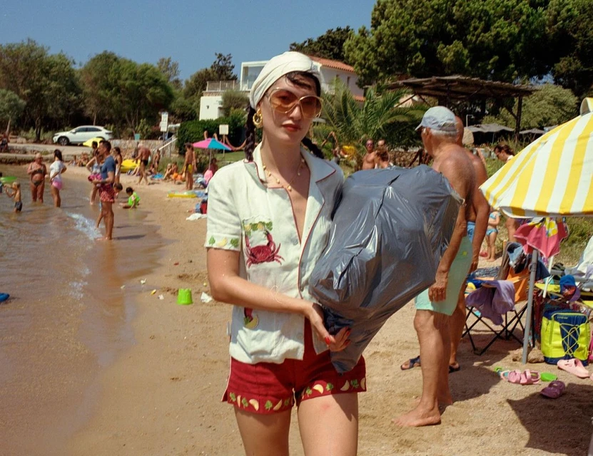 Woman wearing sunglasses on the beach carries a full black garbage bag with people in the water and sand in the background.
