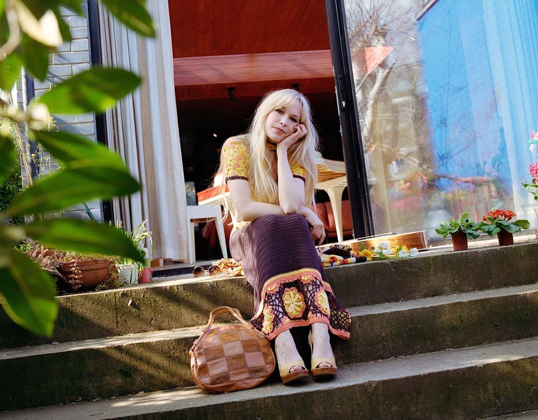 Woman with long blonde hair sits on sunlit steps in a brown crochet skirt and yellow top with a patchwork leather bag, looking relaxed.