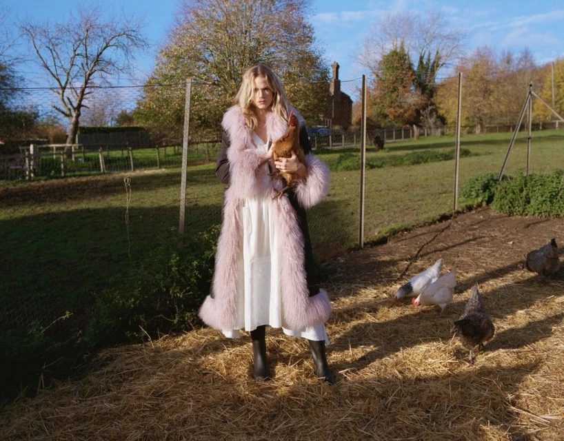 A fashionable woman in a furry coat is holding a chicken in a farm environment surrounded by other chickens and a grassy landscape.