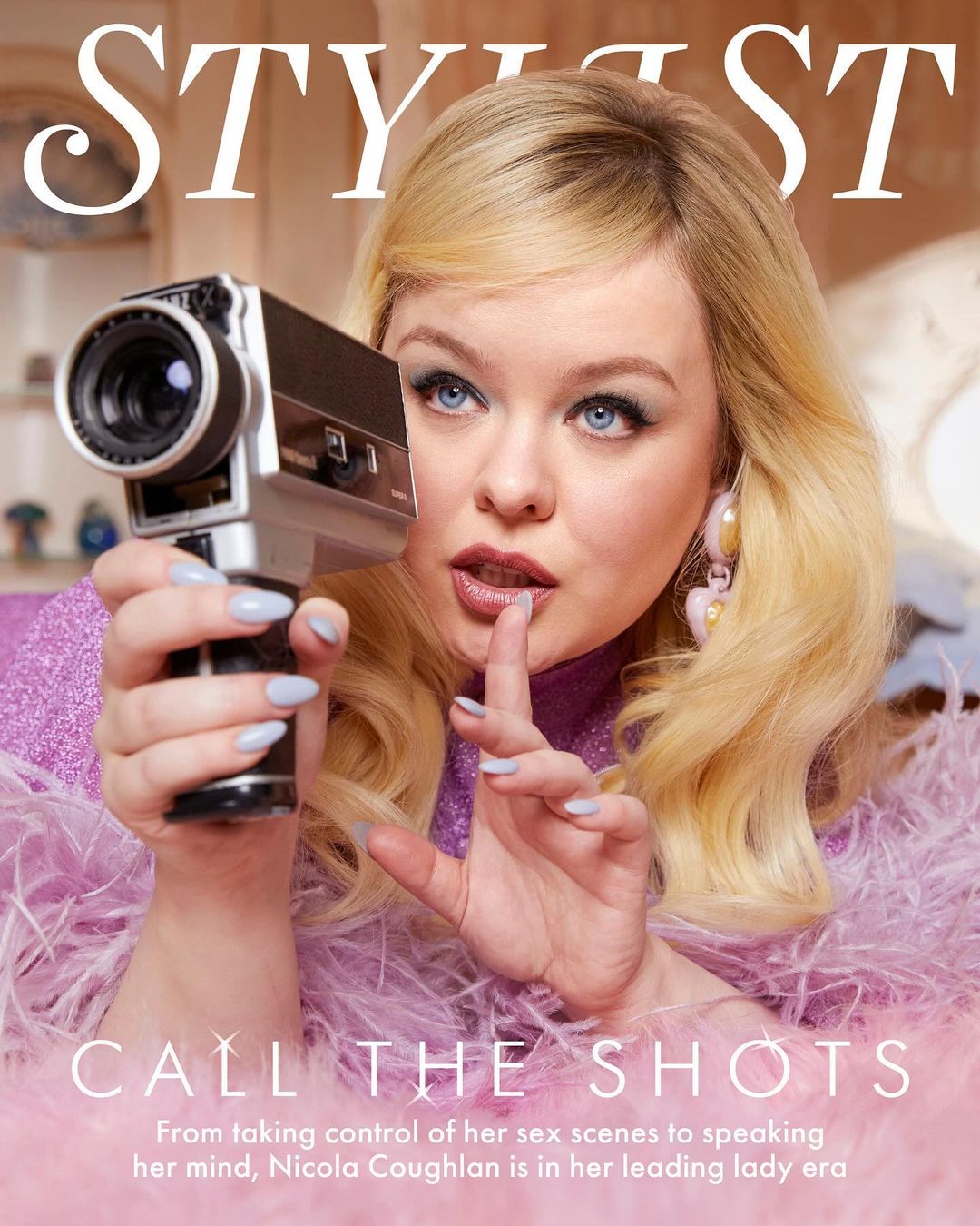 Nicola Coughlan cover shot, holding an old-fashioned film camera in a photo studio with a pink feather boa and blonde hair.