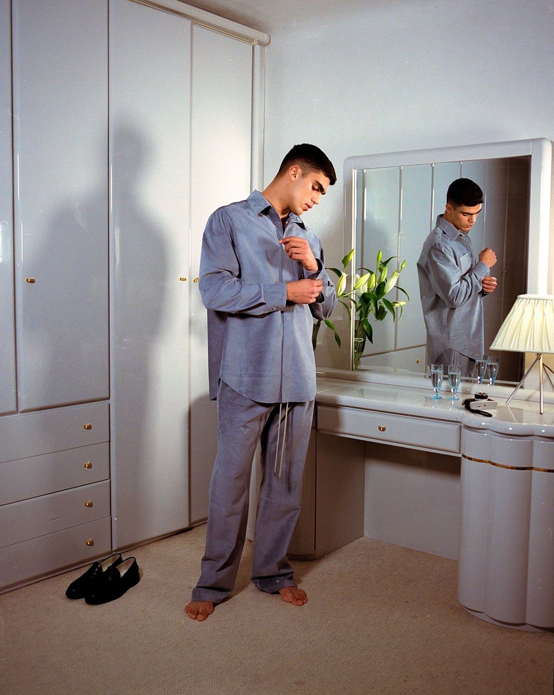 Young adult male in a grey shirt and pants stands in front of a vanity with a mirror, adjusting his cuff in a bright, neutral-toned room.