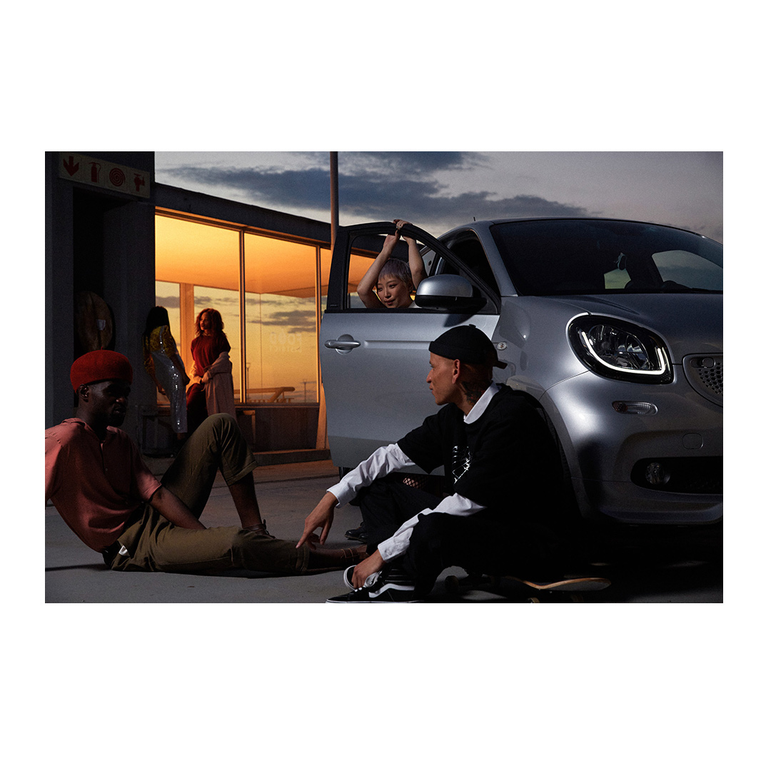 Group of young adults gather by a silver car at dusk, with sunset colors reflecting in the building's windows.