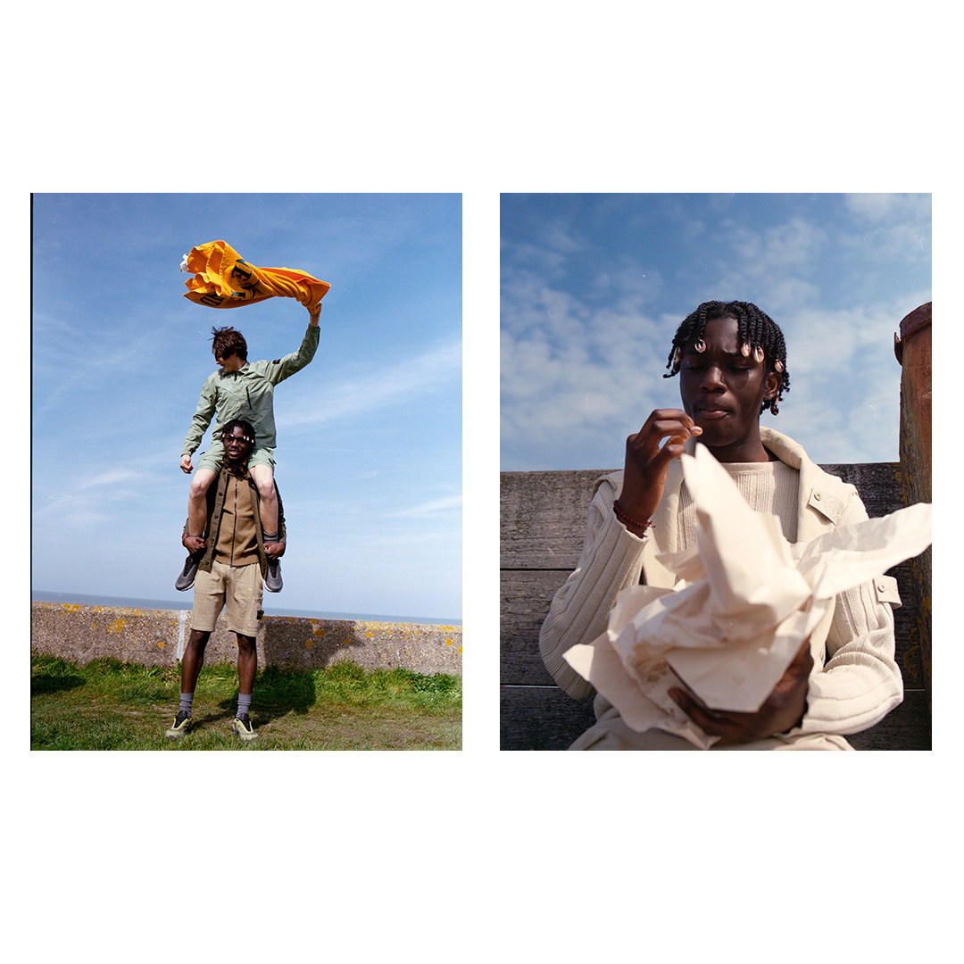 Two dark-skinned men outdoors; one carries the other on his shoulders while the man on top holds up yellow fabric against blue sky.