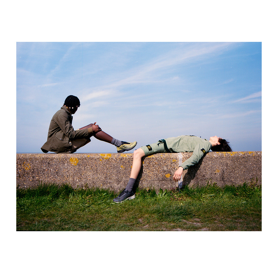 Two people in casual wear sit on a concrete wall against a blue sky backdrop, with one person reclining and the other sitting up.