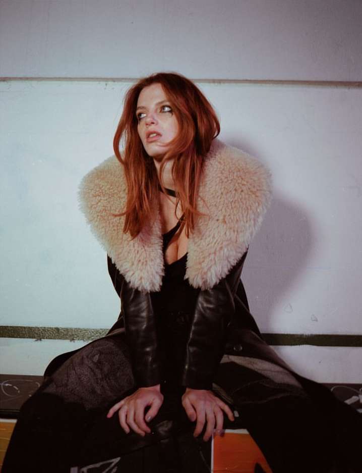 Portrait of a red-haired woman wearing a dark leather coat with a large faux fur collar, posing against a white wall indoors.
