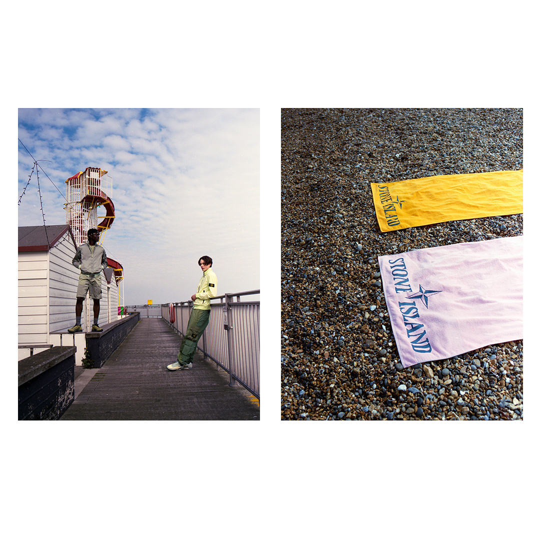 Two stylish men on a boardwalk with a distant seaside structure, alongside a beach with yellow and pink Stone Island branded towels.