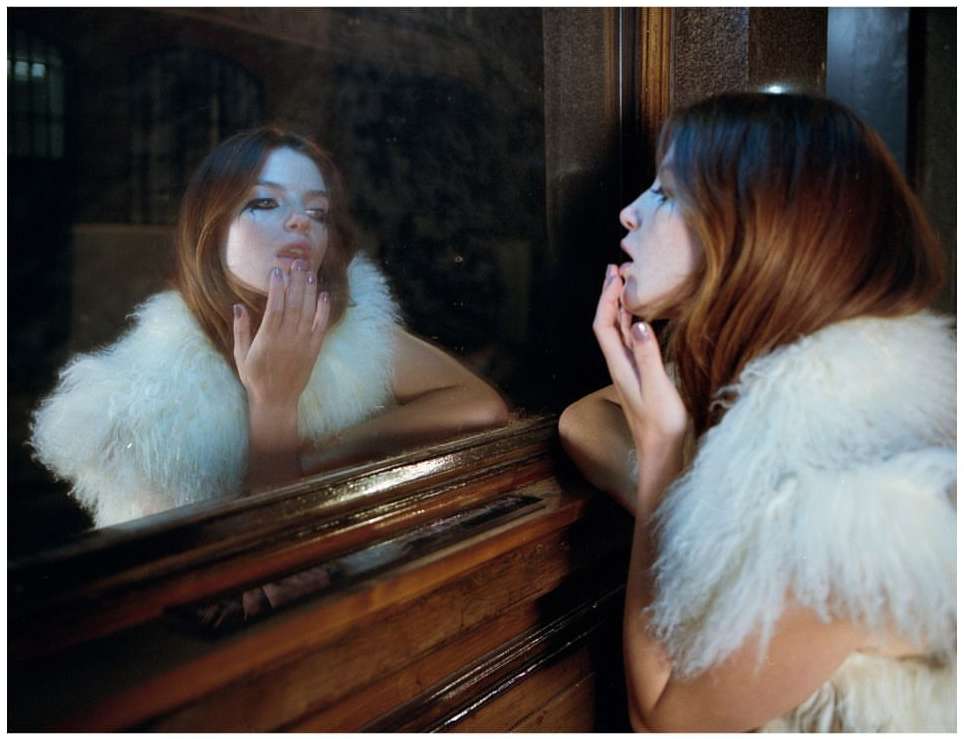Portrait of a woman with red hair and white fur collar looking at herself in the mirror with a dark background, applying lipstick.