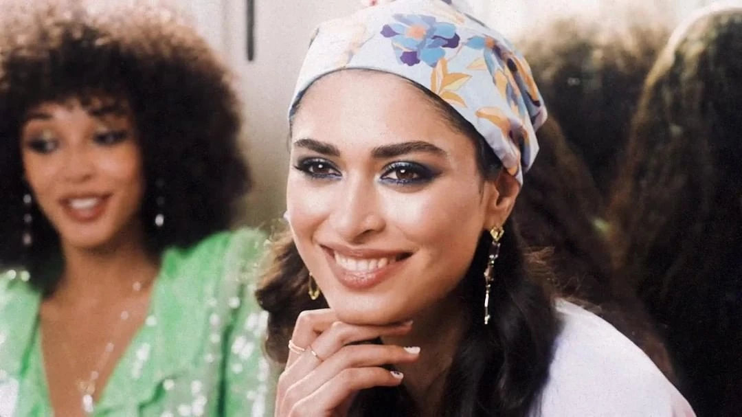 Smiling woman with floral headscarf and hand on chin, accompanied by a second woman in sequined green, indoor setting.