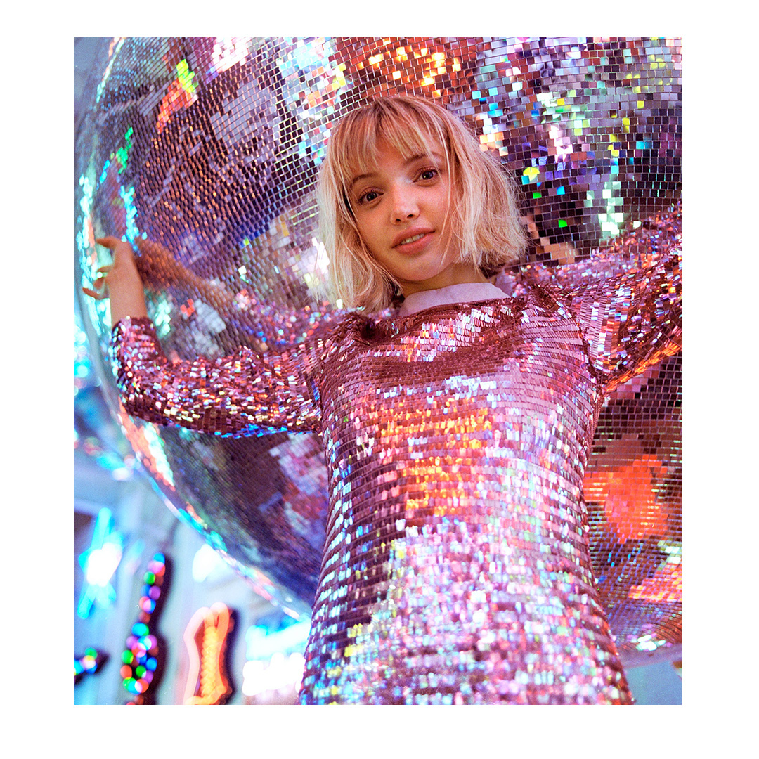 Young woman poses in a shiny pink sequin dress with a large disco ball in a portrait shot with soft lighting and colorful bokeh.