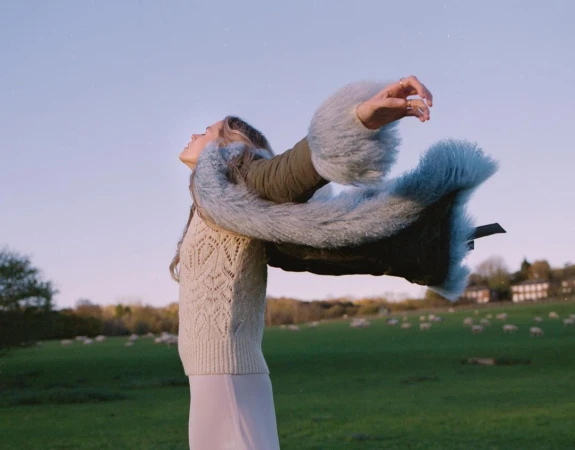 Woman in sweater and fur-trimmed coat with arms raised, gazing upwards in outdoor landscape setting during daytime under a clear sky.