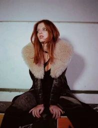 Red-haired woman in a black leather coat with a large fur collar seated against a white wall.