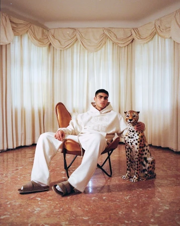 Young man in cream hoodie and sweatpants seated in a leather chair beside a toy leopard figurine