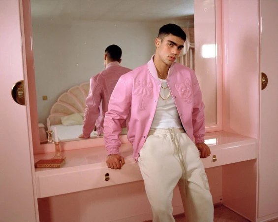Young man wearing a pink bomber jacket and white sweatpants leaning on a matching pink vanity table in a pink room