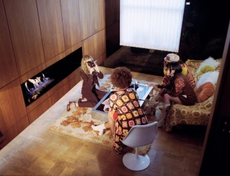 Four women gathered in a retro wood-paneled living room with a modern linear fireplace, photographed from a high angle.