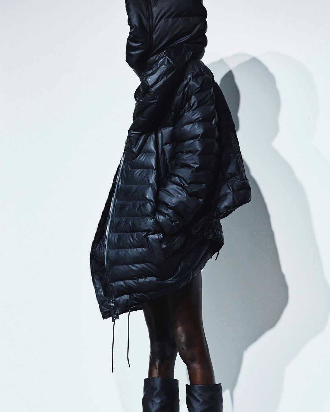 A woman with dark skin wears a shiny black puffer coat with a hood and black boots against a bright white background in a fashion shot.