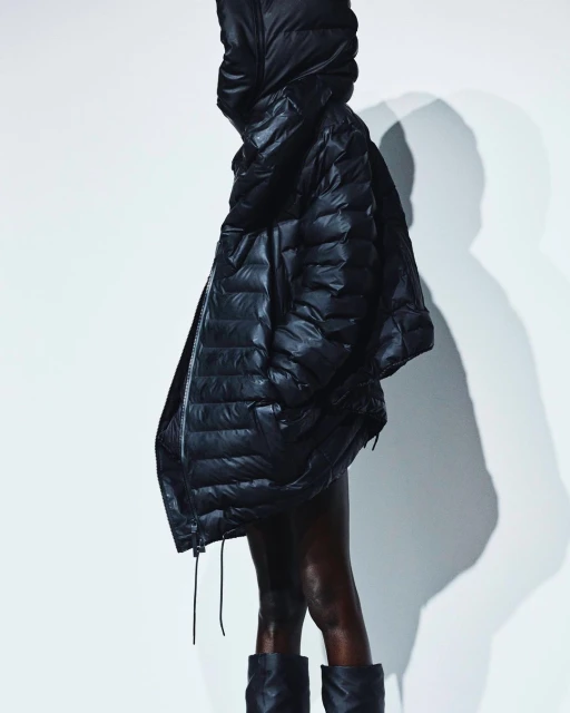 A woman with dark skin wears a shiny black puffer coat with a hood and black boots against a bright white background in a fashion shot.