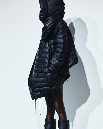 A woman with dark skin wears a shiny black puffer coat with a hood and black boots against a bright white background in a fashion shot.
