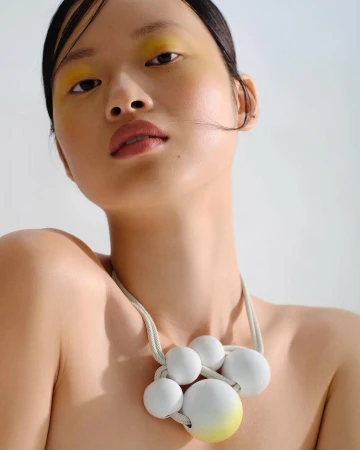 Portrait of a woman wearing a white spherical beaded statement necklace with yellow eyeshadow against a neutral backdrop