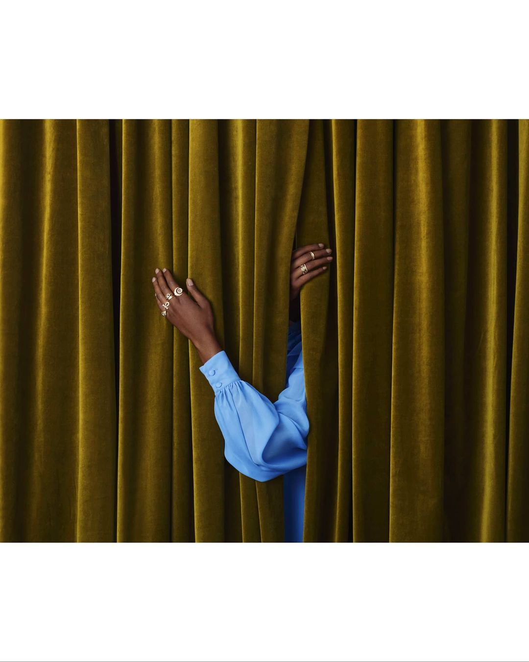 Close-up of a person's hands adorned with rings, peeking through mustard-yellow velvet curtains, creating a sense of mystery and allure.