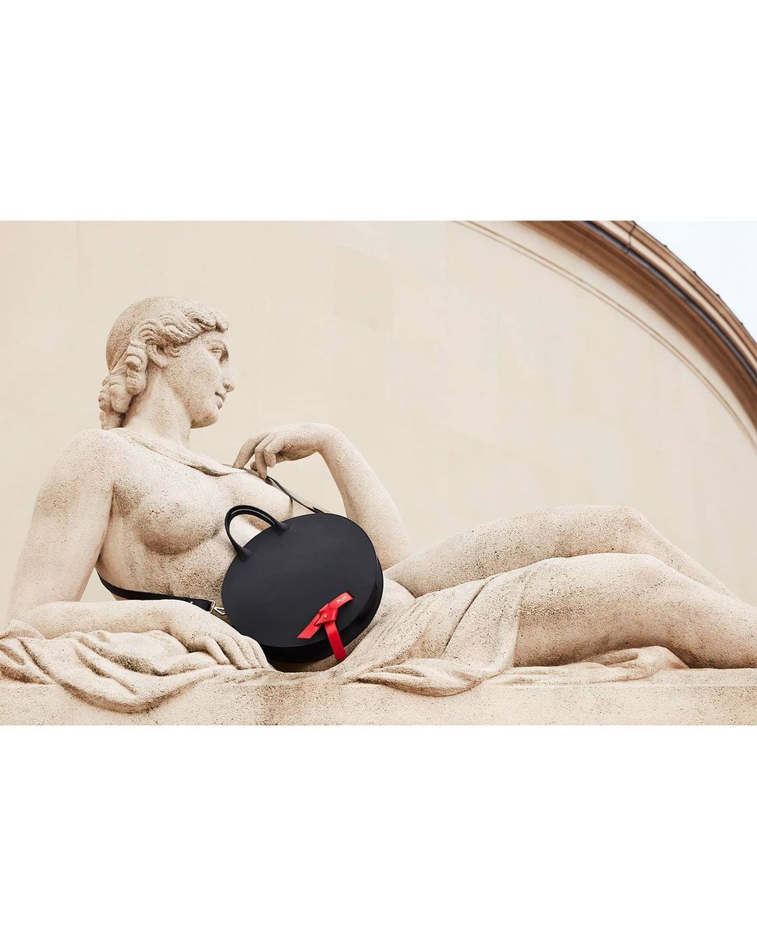 Black, round handbag with a red ribbon is propped against an outdoor sculpture of a reclining human figure in neutral tones.
