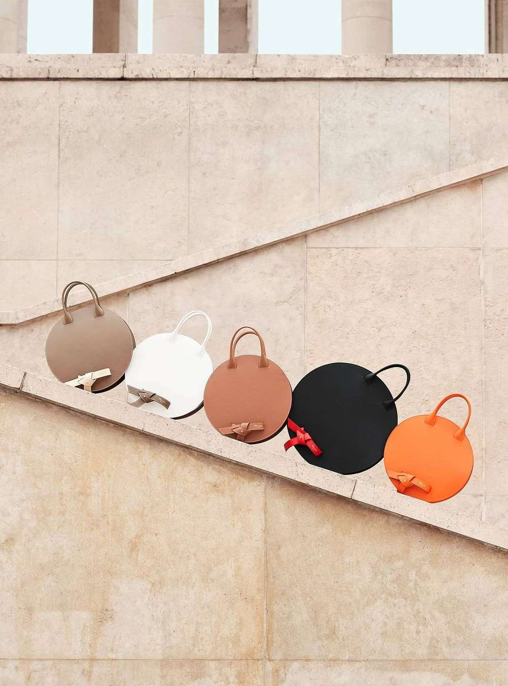 Five round handbags in beige, white, brown, black, and orange with bow details displayed on stone steps with architectural columns.