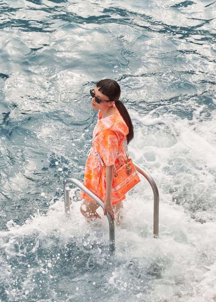 Woman in an orange outfit and sunglasses standing on metal steps in water, holding a handbag with a branded logo in daylight.