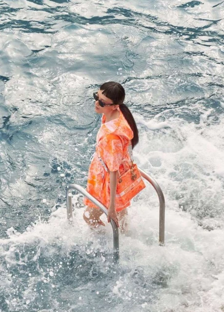 Woman in an orange outfit and sunglasses standing on metal steps in water, holding a handbag with a branded logo in daylight.