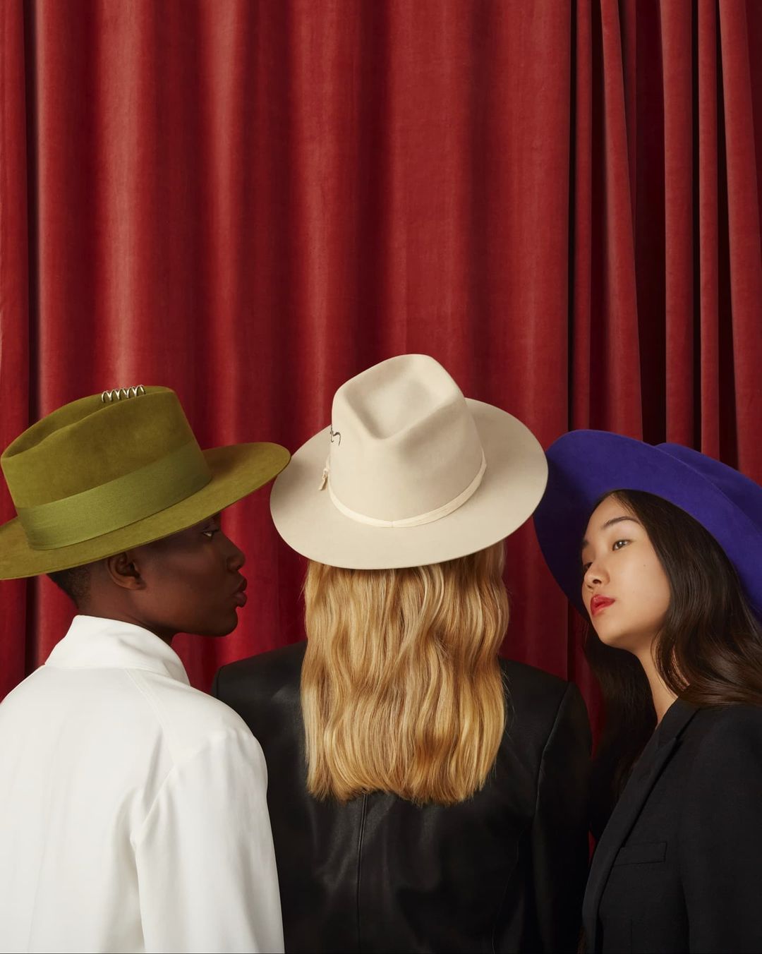 Three people wearing hats of different colors stand in front of a red curtain backdrop. The hats are olive, cream, and violet.