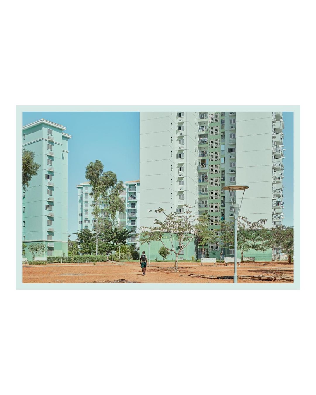 An urban scene shows a person on a bicycle riding across a dirt patch with mid-rise apartment buildings in the background, under a blue sky.