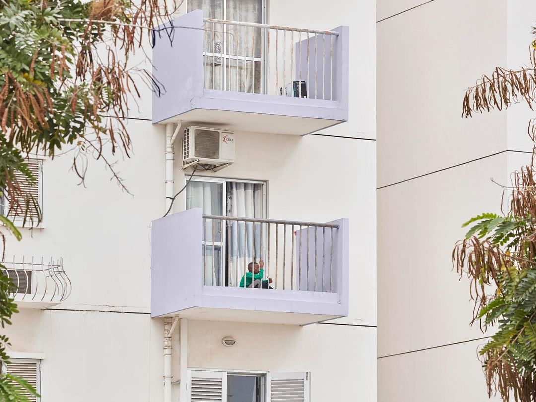 Exterior shot of a modern white apartment building with balconies and a person sitting on one of the balconies, surrounded by green trees.