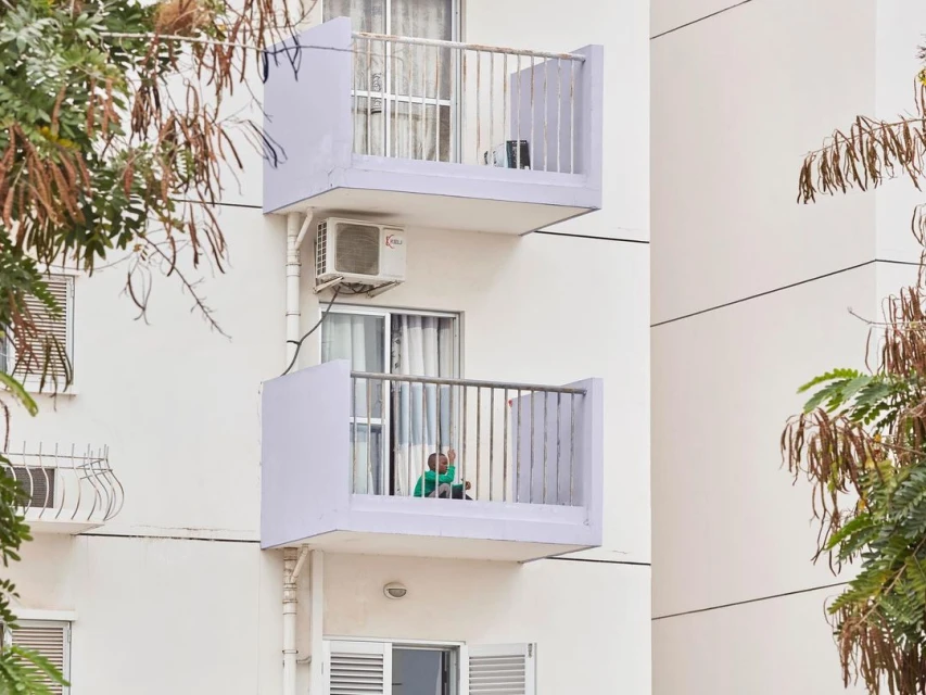 Exterior shot of a modern white apartment building with balconies and a person sitting on one of the balconies, surrounded by green trees.