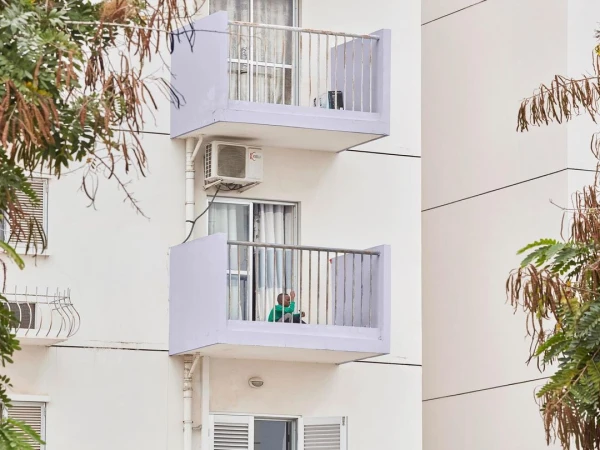 Exterior shot of a modern white apartment building with balconies and a person sitting on one of the balconies, surrounded by green trees.