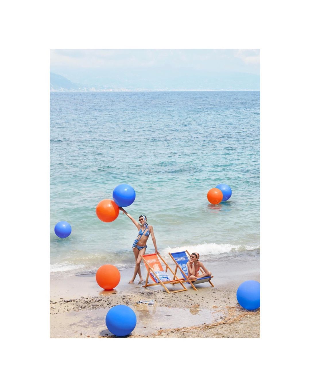 Two women pose among colorful balloons on a sandy beach with turquoise water. One woman holds orange and blue balloons. Sunny day.
