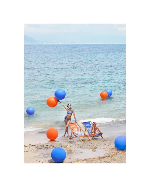 Two women pose among colorful balloons on a sandy beach with turquoise water. One woman holds orange and blue balloons. Sunny day.