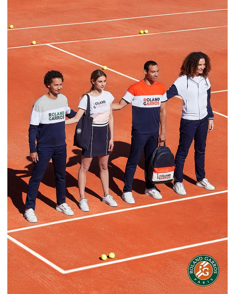 Four adults model Roland Garros tennis apparel on a red clay court, showcasing a coordinated collection in shades of navy, white, and orange.