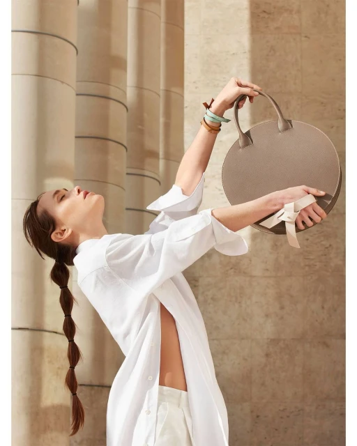 A woman in a white linen shirt holds a round handbag outdoors, posing against a background of cylindrical stone pillars in soft light.