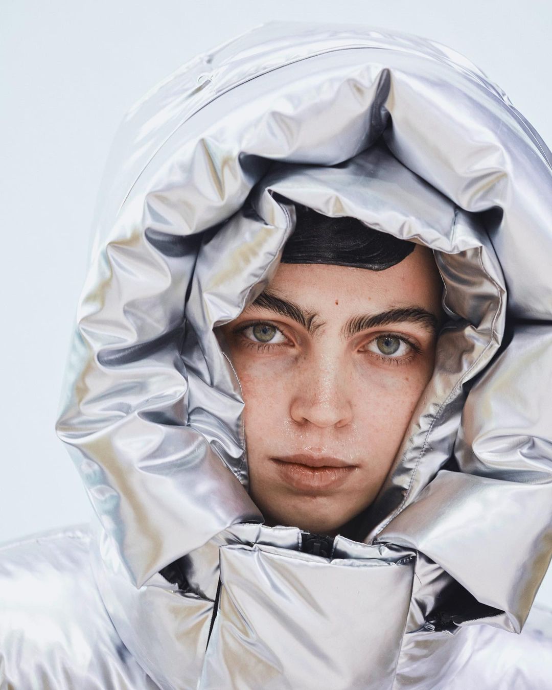 Close-up of a model with green eyes wearing a silver puffer jacket with a hood framing the face against a bright white background.