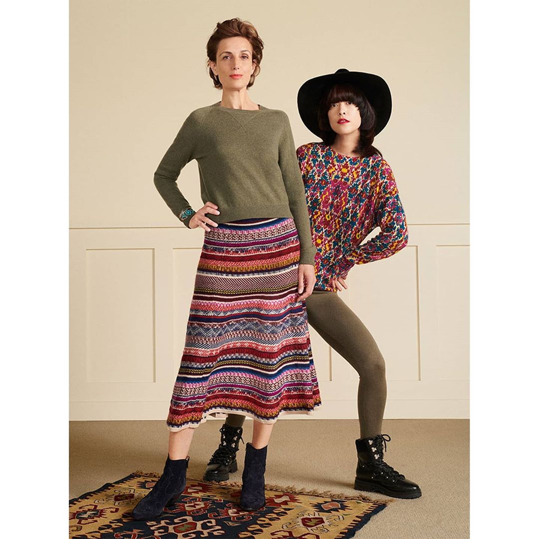 Two adult women pose indoors wearing colorful patterned sweaters and skirts. A neutral wall and patterned carpet complete the fashion shot.