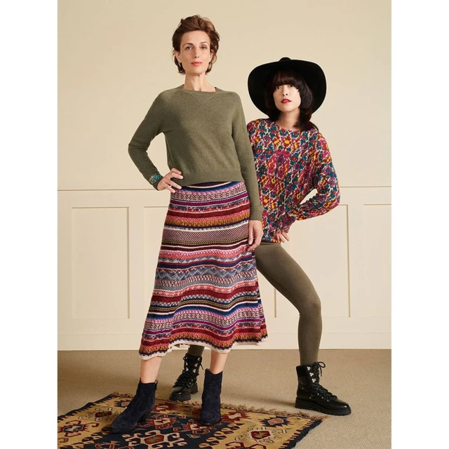Two adult women pose indoors wearing colorful patterned sweaters and skirts. A neutral wall and patterned carpet complete the fashion shot.
