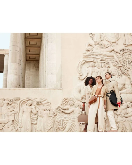 Three women pose with purses against an ornate, beige wall. The women wear neutral-toned clothing. One woman holds a round, black purse.