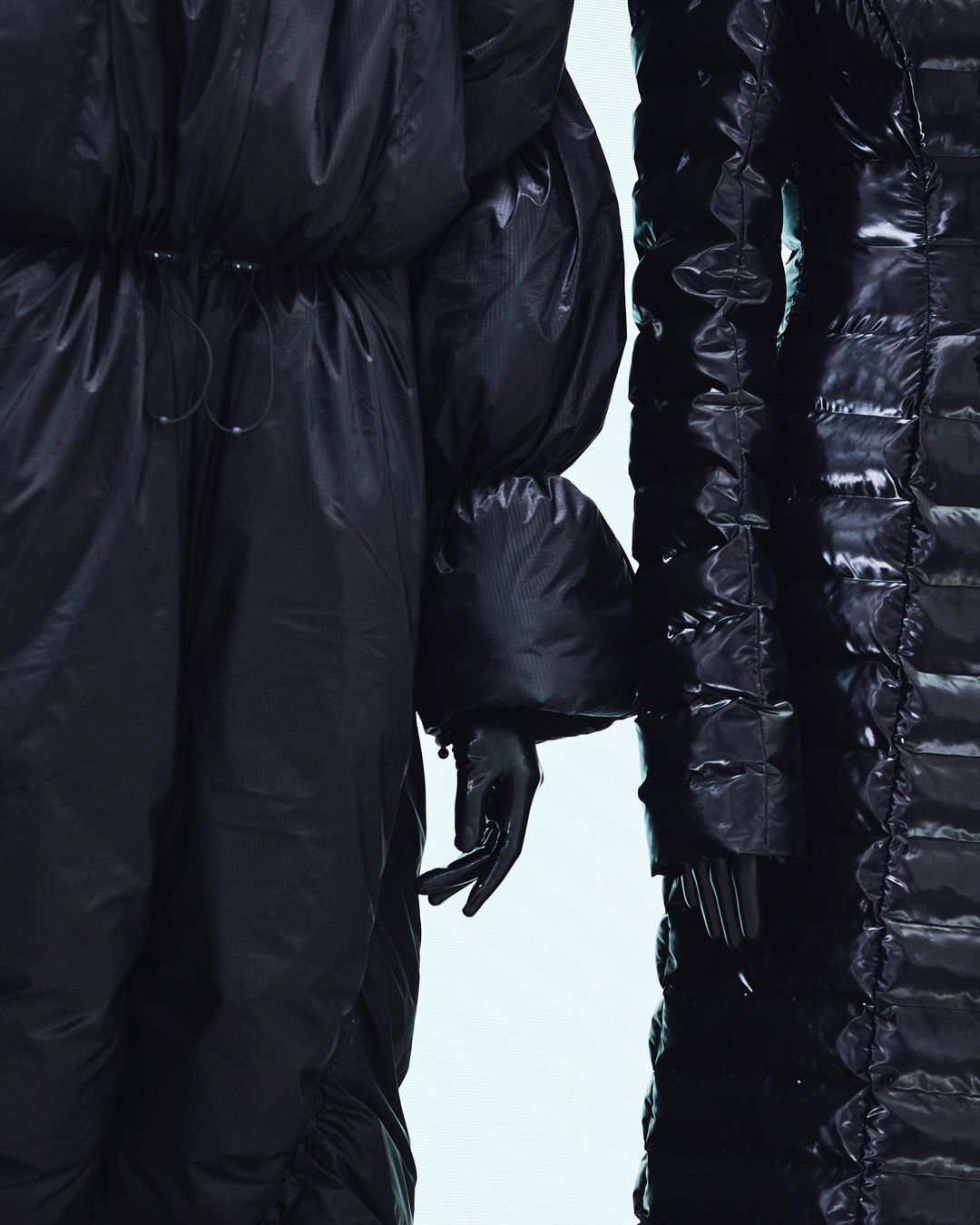 Close-up of two figures in black, shiny puffer coats with detailed stitching in a bright studio setting, fashion forward editorial.