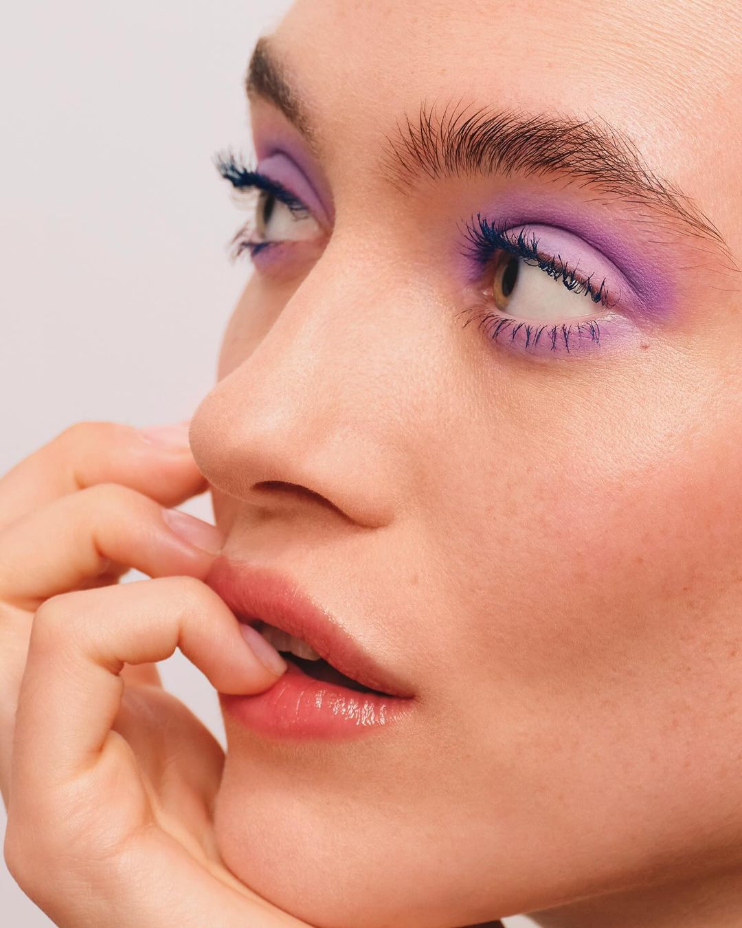 Close-up of a woman's face with purple eyeshadow and blue mascara, pale skin, and fingers near her mouth, against a light background.