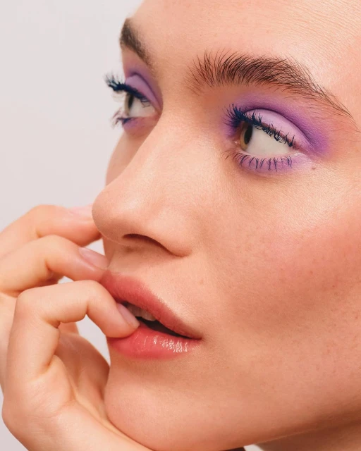 Close-up of a woman's face with purple eyeshadow and blue mascara, pale skin, and fingers near her mouth, against a light background.