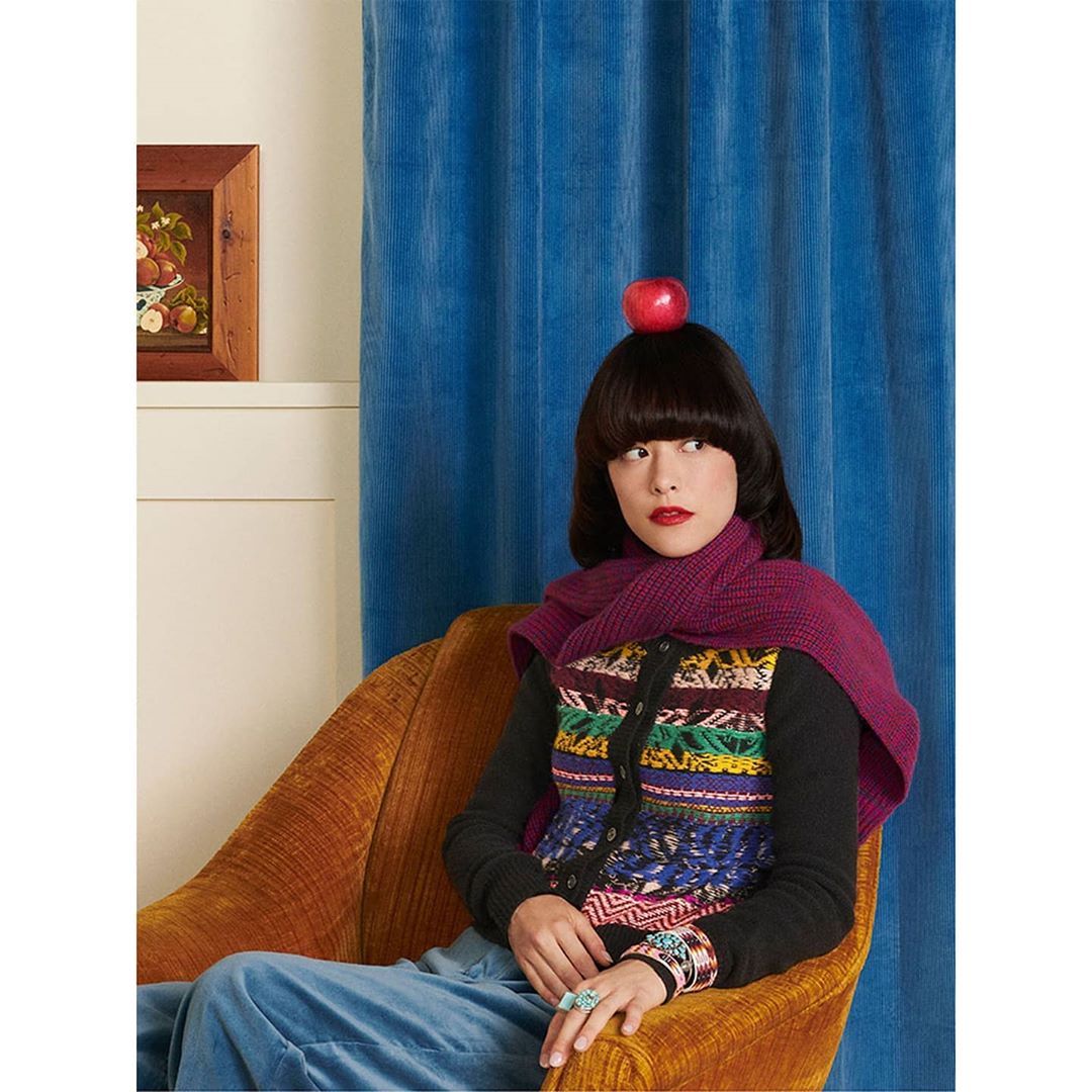 Adult female with bob haircut poses in armchair with apple on head in colorful cardigan, in indoor setting with blue curtains.