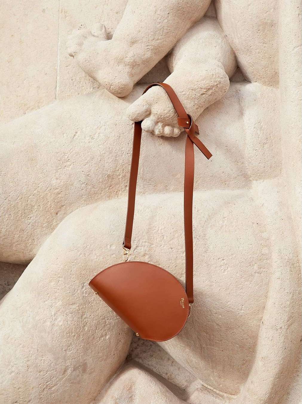 Brown leather handbag with long straps held against a beige stone sculpture, creating a high-fashion editorial product shot.