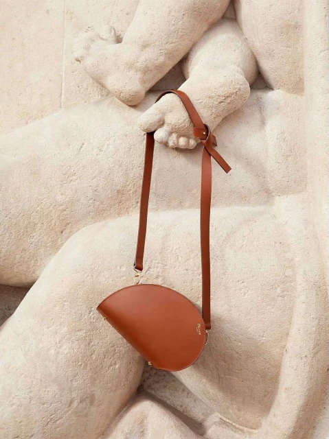 Brown leather handbag with long straps held against a beige stone sculpture, creating a high-fashion editorial product shot.