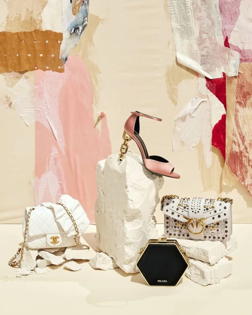 Still life shot of a pink high heel, Chanel bag, studded shoulder bag, and Prada clutch arranged on stacked stone blocks, with painted backdrop.