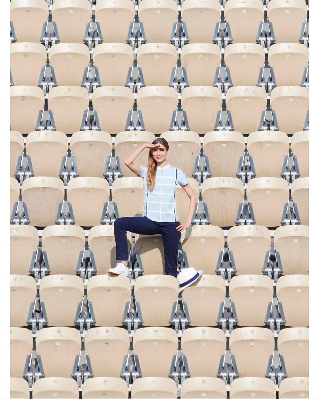 Woman poses in stadium seating, wearing casual attire with a white hat resting on a seat. Rows of empty seats fill the frame.