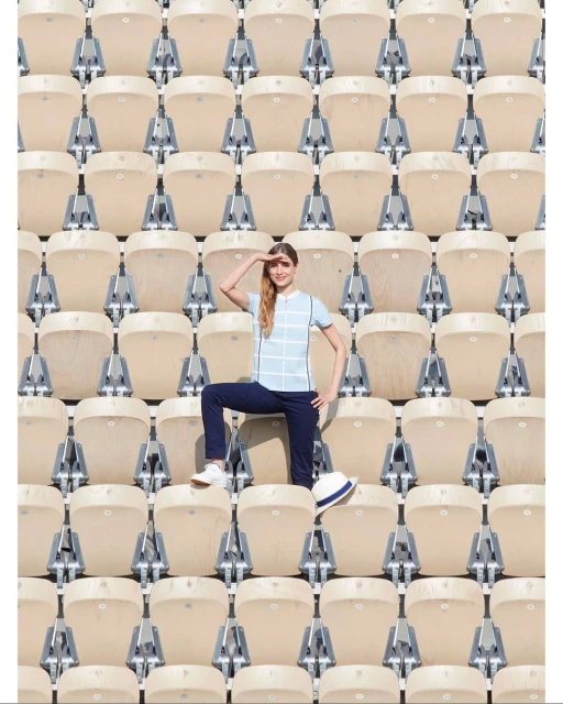 Woman poses in stadium seating, wearing casual attire with a white hat resting on a seat. Rows of empty seats fill the frame.