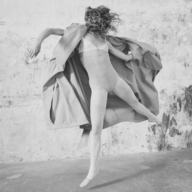 Woman in motion wearing a long coat and fishnet tights, captured in a studio against a textured wall, creating a dynamic black and white fashion portrait.